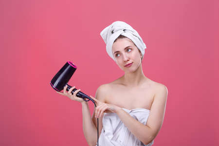 Beautiful girl in a white towel on her head, holds a hair dryer and looks at the stann.の写真素材