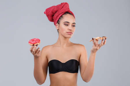 A young girl in a bra and a towel on her head, holds an slice of pizza and a donut, preferring pizza.の写真素材