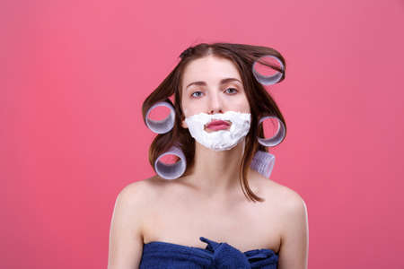 European girl with curlers on hair, shaving foam on face and a bath towel. Pink background.の写真素材