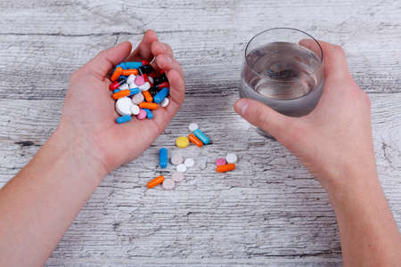 The hands of a European man holding a glass of water and a bunch of different colored pills in capsules and without. Close-up.の写真素材