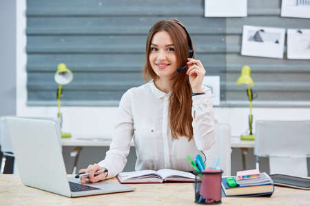 Girl office worker sitting at table in headphones, with laptop and smiling.の写真素材