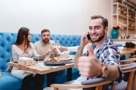Cheerful guy talks on the phone and shows his thumb up, sitting in a cafe with friends.の写真素材