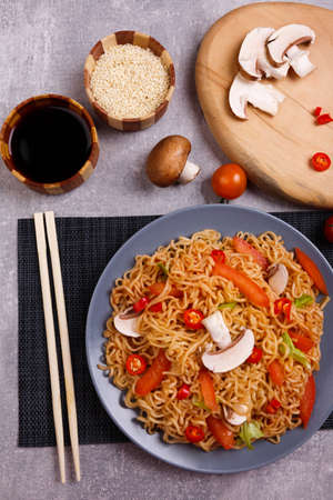 On the table there is a portion of noodles with mushrooms, tomatoes, chili and green peppers, as well as soy sauce and sesame seeds. Top view.の写真素材