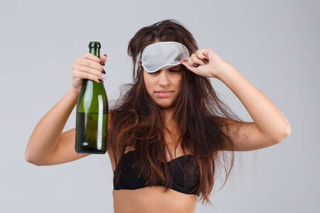 A girl with a bad appearance holds a bottle of alcohol and corrects a mask for sleeping. On a gray background.の写真素材