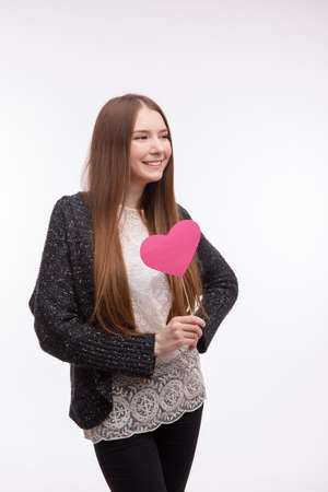 Pretty girl holding a pink heart on a stick, smiling and looking away. Isolated on white.の写真素材