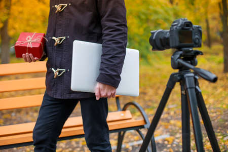A European guy stands on the street in an autumn park in front of a camera on a tripod and holds a red gift box and a gray modern tablet in his hands.の写真素材