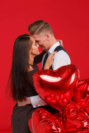Happy couple in love hug each other sensually and smile, next to heart shaped balls. Red background.の写真素材