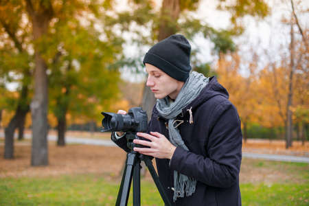A serious young guy, stands outside and sets up a modern camera on a tripod.の写真素材