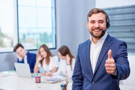 Happy male office worker in suit and headphones stands, smiling cheerfully and showing thumb up.の写真素材