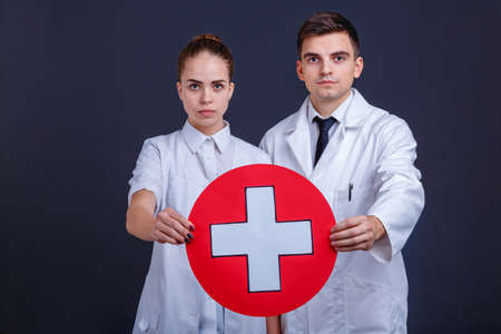Two doctors in uniform, hold one medical sign, a white cross in a red circle.の写真素材
