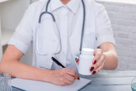 Hands of a girl doctor in uniform sits at the desk and holds a jar with a drug.の写真素材