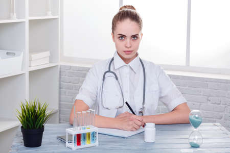 A serious girl, a doctor in uniform, sits at her desk and writes something in a notebook. Indoors.の写真素材