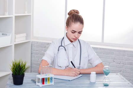 A girl doctor, in white uniform, sits at her desk and writes something to her notebook.の写真素材