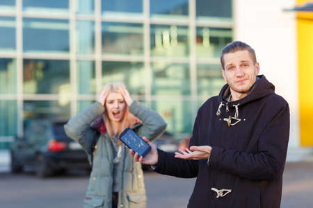 A frustrated guy is holding a smartphone with a broken screen, in the background a girl screams and clings to her head.の写真素材