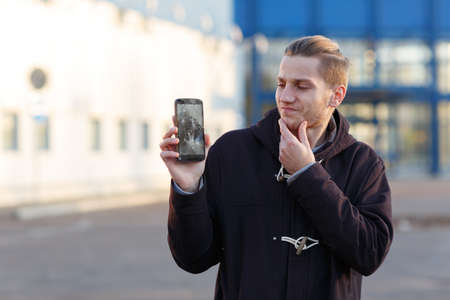 A young guy, holding a mobile phone with a broken screen, frustratedly rubbing his chin.の写真素材