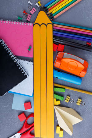 A large yellow paper pencil lies on scattered different colored pencils, several notebooks, clamps...