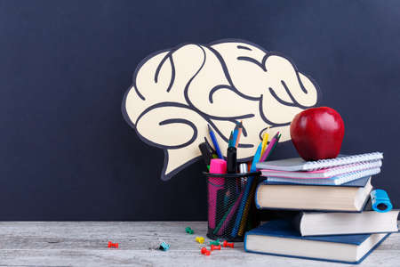 A small pile of books and notebooks, various stationery and a red juicy apple, on a dark background with a picture of the brain. The concept of education.の写真素材