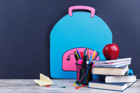 A small stack of books and notebooks, various stationery and a red juicy apple, on a dark background with an image of a paper backpack. The concept of education.の写真素材