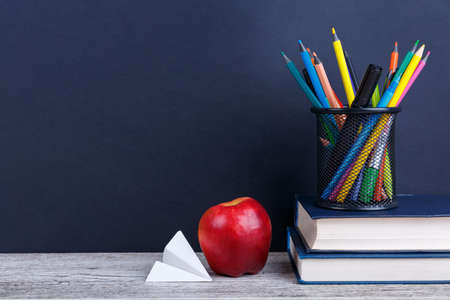 A small stack of two books, a red juicy apple, colorful pencils in a black mesh case and a paper...