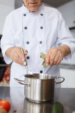 The elderly chef is cooking a soup. Grandfather with a metal spoon and pan adding a laurel leaf to...