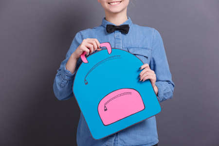 A young girl in a blue shirt with a big paper backpack on a gray blackboard. School objects conceptの写真素材