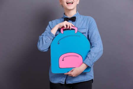 A young girl in a blue shirt with a big paper backpack on a gray blackboard. School objects conceptの写真素材