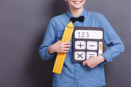 A young woman holds an image of a yellow pencil and a calculator on a gray background. Objects for work with documents.の写真素材
