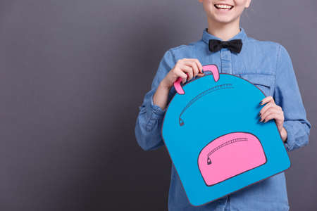 A young girl in a blue shirt with a big paper backpack on a gray blackboard. School objects conceptの写真素材
