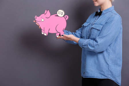 A young woman holds a big paper pink piggy bank with a coin on a gray background. The concept of money and financeの写真素材