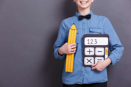 A young woman holds an image of a yellow pencil and a calculator on a gray background. Objects for work with documents.の写真素材