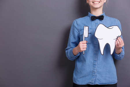 A young woman in blue shirt holds a paper image of a big paper tooth and a toothbrush on a gray background. Healthcare concept.の写真素材