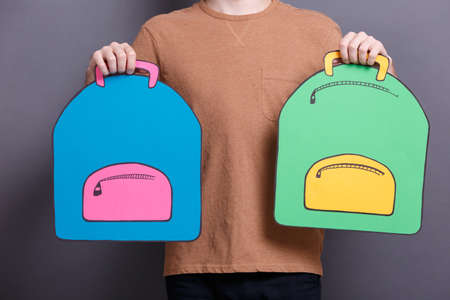 A young guy holding two colorful paper backpack on a gray background. The concept of education and school objectsの写真素材