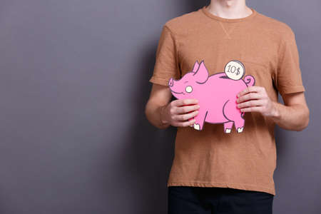 A young man holds a big paper pink piggy bank with a coin on a gray background. The concept of money and financeの写真素材
