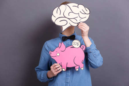 A young woman holds a big paper pink piggy bank with a coin and a big brain on a gray background. The concept of money and financeの写真素材