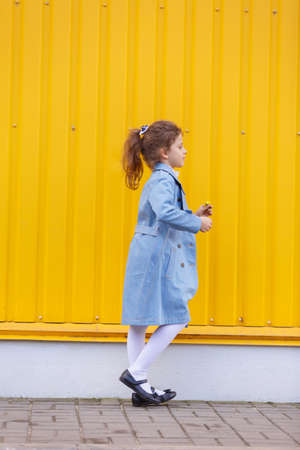 A cute little girl in a blue denim coat on a bright yellow background outdoors. Fall fashion clothes for kids concept.の写真素材