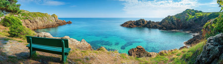 Panoramic view of the coast and cliffs at Guernsey on a clear day, with an empty green bench overlooking calm turquoise waters and lush coastal vegetation under a clear blue sky. The picturesque scene captures the vibrant colors of nature against a backdrop of rugged coastline. This photograph highlights the beauty and tranquility found on the Channel Islands. --ar 52:15 --v 6.1 Job ID: 8128aa21-d883-4b50-83be-e7bc1755b1b6の素材