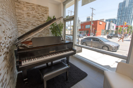 A grand piano sits in a well-lit room next to a large window offering a cityscape view. The piano, with its open lid and keys, is the focal point, complemented by a piano bench. The room features brick walls and a neutral color scheme. This scene could be used for musical or educational content.の素材