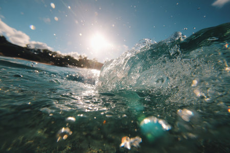 An ocean wave is captured mid-crest, showcasing transparent water illuminated by strong sunlight. The composition emphasizes the dynamic movement of the water with sprays and bubbles. A blue sky forms the backdrop, hinting at an outdoor, daytime environment. Suitable for editorial and commercial applications.の素材
