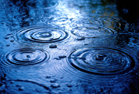 An overhead shot captures the impact of raindrops on a water surface, generating concentric circles. The image displays varying shades of blue, with light reflecting off the ripples. This photograph could be utilized in design projects, illustrative backgrounds, or for conveying concepts such as change or impact.の素材