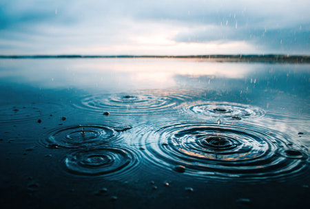 The image features water surface marked by concentric ripples from raindrops. The water reflects a gray cloudy sky. The composition emphasizes depth and texture with the water in the foreground and a hazy horizon. Suitable for atmospheric visuals or representing environmental themes.の素材