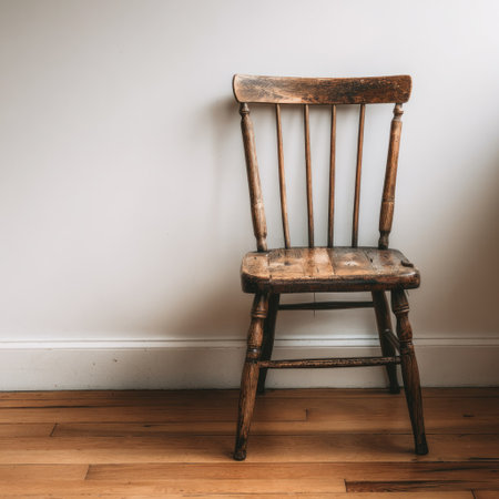 An aged wooden chair stands against a simple white wall, illuminated by natural light. The chair exhibits a weathered texture, showcasing the grains of the wood. The scene is completed by a hardwood floor, suggesting an indoor domestic setting. This image could be suitable for various editorial and commercial applications.の素材
