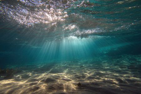 An underwater scene depicts sunlight filtering through the ocean surface. The clear water reveals a sandy sea floor with ripples. The composition highlights the interaction of light and water, creating a serene and captivating effect. Suitable for use in various visual media, including editorial and commercial projects.の素材