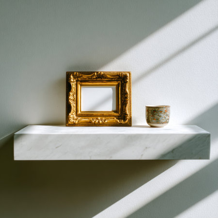 A square frame with ornate gold details and a small decorative cup sit on a white shelf against a pale wall. The scene is bathed in contrasting light and shadow, highlighting textures. The composition offers a minimalist aesthetic suitable for various design, art, or lifestyle related commercial uses.の素材