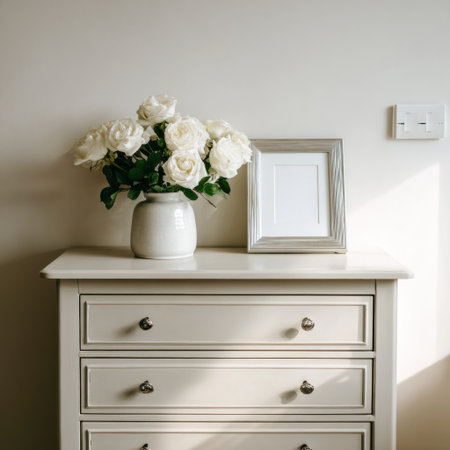 A close-up captures a bouquet of white roses in a ceramic vase atop a dresser. A framed picture rests beside the flowers. The scene is illuminated by natural light, creating soft shadows. The composition suggests a serene indoor setting with potential uses in interior design, or lifestyle content.の素材