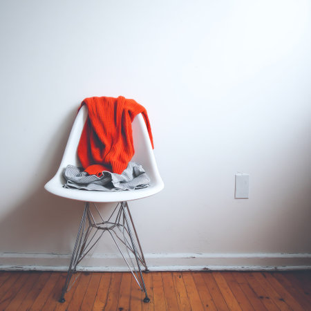 A white chair with a bright orange garment draped over it is positioned against a plain white wall. The composition features wooden flooring and a wall outlet adding to the simplicity. The lighting is diffused creating soft shadows. Suitable for use in design concepts, editorial projects, or lifestyle illustrations.の素材