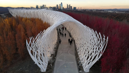 An overhead shot reveals a white, intricate bridge design extending over colorful trees. Silhouetted figures traverse the walkway. The scene displays an outdoor setting with warm autumnal hues. The image suggests potential for commercial projects and editorial content related to architecture and design.の素材