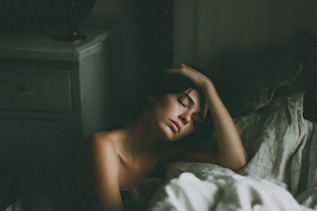 A woman reclines, eyes closed, bare shoulder exposed, resting on a bed in soft lightの素材