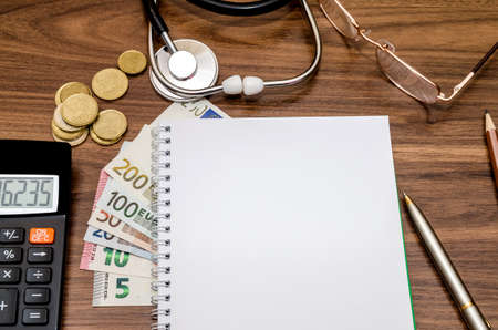 empry notepad with stethoscope, euro money, glasses, pen, calculator on table