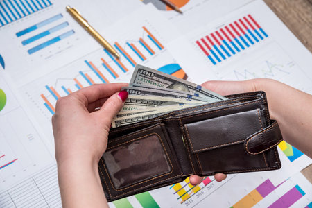 female hands holding leather wallet with US Dollars cash inside above business charts