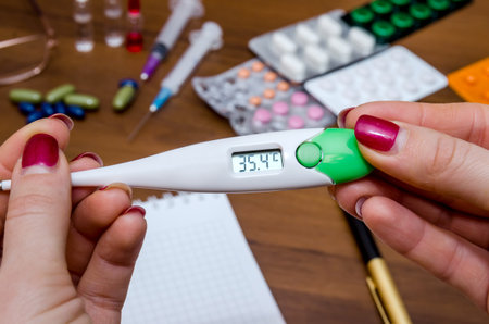 Hand holding thermometer above pills, syringe, tablet, notebook and pen